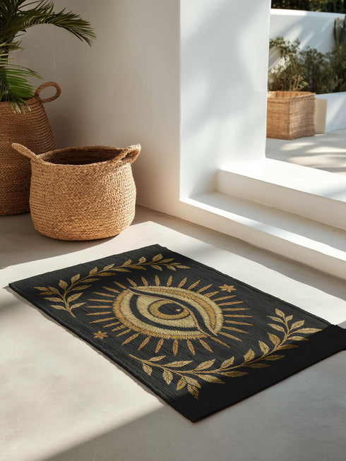 Woven 24x36 inch evil eye doormat in black and gold, featuring a central embroidered eye surrounded by sun rays and olive branches, made of 100% polyester chenille with a non-slip backing, designed for mystical or gothic home decor.